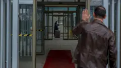 Deri ceketli bir adam kapıların ardındaki bir kadına el sallıyor. Kadın, elinde valize adama bakıyor. 