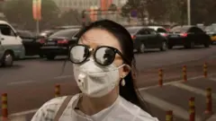 Seorang perempuan berjalan di trotoar kota sambil mengenakan kacamata dan masker.