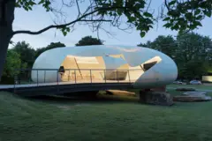 Pabellón contemporáneo de forma orgánica, iluminado desde el interior y elevado sobre una plataforma metálica en un parque.