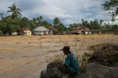 বন্যার স্রোতের পাশে বসে আছেন একজন পুরুষ, অন্যপাড়ে কয়েকটি ঘর দেখা যাচ্ছে