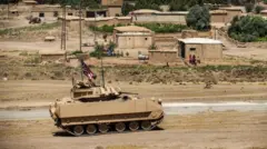 Üzerinde ABD bayrağı olan çöl kamuflajlı bir zırhlı araç yolda ilerliyor. Arkada tek katlı binalar ve bir grup kadın ve çocuk var. 