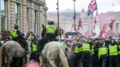 Конные и пешие полицейские в светоотражающих жилетах пытаются удержать толпу с флагами Англии и Соединенного Королевства на стыке Уайтхолла и Трафальгарской площади в Лондоне под дождем