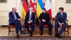 男性4人が横並びで椅子に座っている。背後には、クリーム色に塗られた木製の壁と大理石の暖炉がある。足元には大きなじゅうたんが敷かれている 