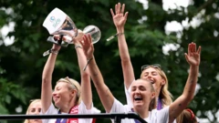 Девушки в белом подняли руки вверх, одна из них держит кубок. У всех радостные лица