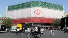 エンケラブ広場に臨む建物の二つの壁面にイラン国旗が大きく掲示されている。その前を車両や歩行者が行きかう