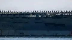 На палубі великого авіаносця вишикувалися десятки моряків. Корабель стоїть біля причалу, а його масивний корпус займає майже весь кадр.