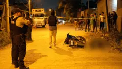 Policiais de pé ao lado de um corpo e de uma motocicleta virada na rua, com pessoas observando atrás da fita da polícia