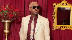 Pretty Mike kootu oyinbo alawọ miliki, o wọ shaat alawọ waini, o si lo tai alaranbara, o fi ọda goolu kun irun rẹ, o lo igo dudu soju, o si ki ọwọ ọtun bọ apo rẹ. Asọ alawọ waini lo wa lẹyin rẹ, gilaasi kan wa lara ogiri ni ọwọ alaafia rẹ, igi kan to gbe ododo duro si wa ni apa ọtun.