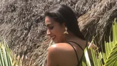 Catalina, entre ramos de palmeira, olha para o lado e sorri, com as costas voltadas para a câmera. Ao fundo, um teto de palha