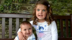 James ve Kate dışarıda ahşap bir bankta oturuyorlar. Kate'in bir kolu James'in omzunda, arka planda ise taş bir duvar ve yeşillikler var.