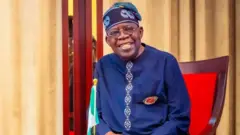 Onyeisiala Naịjirịa bụ Bola Tinubu yi uwe n'acha anụnụ anụnụ ma nọọrọ na ọche nwere akwa uhie uhie nakwa aja aja. Ọkọlọtọ Naịjirịa nọ ya n'ihu dịka ọ na amụrị ọnụ ọchi. O yi okpu n'acha anụnụ anụnụ ma yirikwa igobe anya.