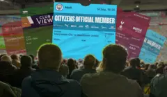 A group of fans are sitting in the stands at a football watch, in front of them are a number of large scale tickets for Manchester City, Everton, West Ham, and Arsenal