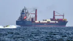 Un barco de carga navega hacia la derecha, mientras una lancha parece acercársele rápidamente.