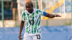 Ngozi Okobi-Okeoghene arbore des cheveux courts teints en blond et porte un maillot de football nigérian blanc et vert ainsi qu'un short blanc floqué du numéro 13. Son bras gauche est tendu sur le côté pour garder l'équilibre tandis qu'elle regarde attentivement le terrain pendant un match de football. À l'arrière-plan, on aperçoit, de façon floue, des barrières placées devant une tribune.