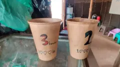 Dua gelas kertas dengan harga untuk tiga jam dan satu jam akses internet, terlihat diletakkan di atas meja di sebuah ruangan sederhana dengan kursi plastik rendah di sepanjang dinding.