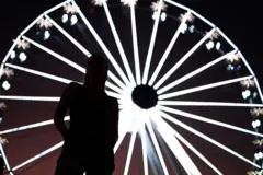 Silhueta de uma pessoa em primeiro plano, de pé e com as mãos atrás do corpo, diante de uma grande roda-gigante iluminada à noite. As luzes brancas da roda formam linhas radiais brilhantes ao fundo, criando um efeito visual circular e contrastando com o corpo escuro da pessoa.