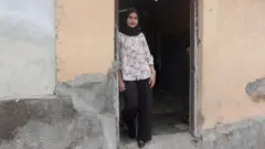 Fatma na porta de sua casa vestindo calças pretas, hijab e uma camisa florida.