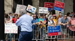 Pessoas seguram cartazes com os dizeres: “Nos Estados Unidos, a lei é o rei!” e “Sem rei! Sem tirania!”, enquanto permanecem atrás de uma barreira metálica