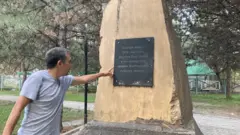 Элери Битикчи Үркүнгө арналган эстеликтеги азыркы жазууну көрсөтүп жатат