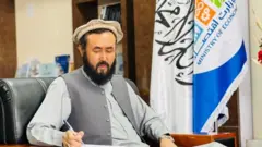 لطیف نظری، معین وزارت اقتصاد حکومت طالبان، در حال نوشتن که در پس زمینه اش پرچم حکومت طالبان و وزارت اقتصاد قرار دارد.