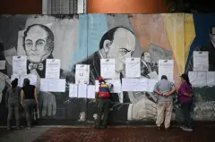 Local de votação na Venezuela em 25/05, com pessoas examinando papéis afixados a uma parede com pinturas