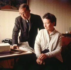 Sartre, de pé, de terno e gravata, com a mão em um jornal sobre a mesa, e ao lado dele está De Beauvoir, sentada, usando um casaco bege e com o cabelo preso.