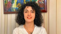 Narges Mohammadi sonríe en esta foto de folleto sin fecha. Tiene el pelo negro rizado y lleva una camisa blanca, un collar en forma de triángulo y lápiz labial rojo.