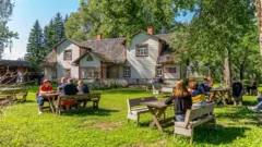 Un grup de persoane stau în jurul unor mese din lemn în fața unei locuințe din Estonia, într-o grădină.