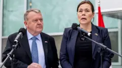 ダークスーツに白いシャツ、青いネクタイを着けた男性が、左隣に立つダークスーツの女性を見ている。女性はブラウンの髪を後ろでまとめ、マイクに向かって話している