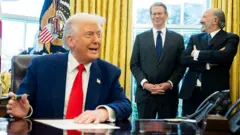 ትራምፕ ፊርማቸው እያኖሮ እና ፈገግ ብለው እየተናገሩ፤ ከኋላ ባለለልጣኖቻቸው እየሳቁ ቆመው ይታያሉ