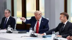 Keir Starmer brit miniszterelnök, Donald Trump amerikai elnök és Emmanuel Macron francia elnök egy fehér asztalnál ülnek. Donald Trump jobb kezét felemeli, és előre felé mutat.