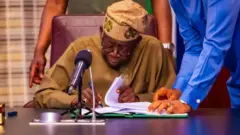 Aarẹ Bola Tinubu lasiko to n buwọ lu iwe nileeṣẹ Aarẹ, o mu gege dani, bẹẹ ni ọwọ kan wa lori tabili rẹ, nigba ti ẹnikan duro lẹyin rẹ.