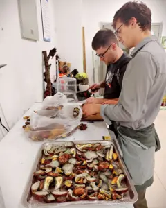 Dos hombres, uno de ellos Domnauer, en una cocina preparan y cortan hongos frescos sobre una mesa con una bandeja llena de hongos rebanados.