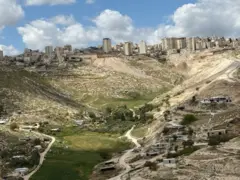 Una agrupación de viviendas beduinas en una zona verde, cerca de la cual se ven colinas con edificios residenciales en sus cimas. La foto fue tomada durante el día.