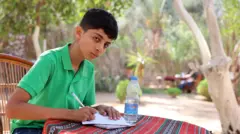Com uma camisa polo verde, ele se dedica às tarefas escolares em uma mesa no jardim. Enquanto escreve no papel com uma caneta, ergue o olhar para a câmera.