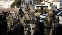 一軒家が並ぶ住宅街に、重装備で顔を隠した迷彩服姿の当局者が複数立っている