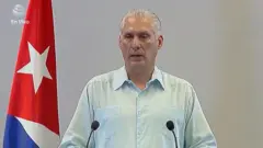 Miguel Díaz-Canel de pie ante un podio. Viste una camisa blanca de manga larga y tiene ambas manos colocadas sobre el podio. Al fondo, al lado izquierdo de la imagen, se ve una bandera de Cuba.