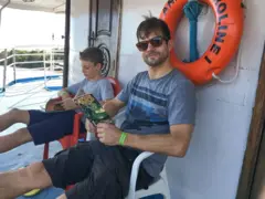 Um homem sentado em uma cadeira, dentro de um barco, lendo um gibi. Ao lado dele há uma criança.