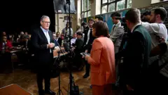 Starmer bir kiliside çok sayıda basın mensubu karşısında duruyor, geride sıralarda oturan resmi giyimli çoğu erkek kişiler var. 