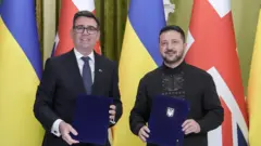 Бернем (ліворуч) і Зеленський (праворуч) у формальному одязі стоять перед прапорами України та Великої Британії. Кожен тримає синю папку з документами. 