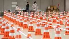 Três mulheres de máscara trabalham em sala dividida por fileiras de cones