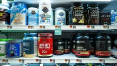 Prateleiras de supermercado com uma variedade de suplementos alimentares, principalmente proteínas em pó, whey protein, colágeno, creatina e produtos à base de plantas.