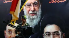 يقف أحد أفراد الجيش الإيراني حارساً أمام صورة المرشد الأعلى الإيراني الراحل آية الله علي خامنئي (أعلى) والمرشد الأعلى الإيراني آية الله مجتبى خامنئي (يمين)

