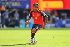 Lamine Yamal durante jogo da Espanha. Ele está chutando a bola e com a camisa vermelha da seleção.
