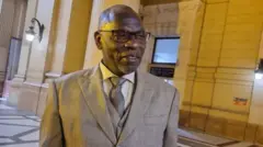 Séraphin Twahirwa, ahagaze amwenyura, yambaye ikoti na 'cravate', inyuma ku ruhande rumwe hari itara ryaka, no ku rundi hari itara ryaka