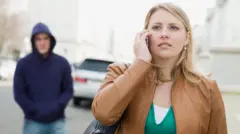 Mulher falando ao celular na rua, com homem encapuzado seguindo atrás