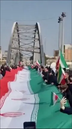 පාලමක අසල දිග ඉරාන කොඩියක් අල්ලාගෙන ජනතාව පෙළ ගැසී සිටී.