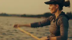 Uma mulher com viseira, brinco de argola e cabelo preso rema enquanto olha para o horizonte.