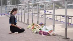 
砂浜を前にした遊歩道に手すりが並び、その前に多数の花束が供えられている。女性がその前に正座をして、両手を広げて手のひらを上に向け、目を閉じている。手すりには、白と青の制止線が張られている