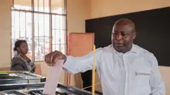 Prezida w’u Burundi Évariste Ndayishimiye ubwo yari arimo gutora mu matora y'abashingamateka, kw'ishure ry’intango rya Musama, i Gitega, kw’itariki ya 5 Ruheshi (6) mu 2025. Yambaye ishati y’umweru, arimo gushyira urupapuro rw’itora mw’isandugu ya plastike y’umukara, mu cumba hahagazemo umugore ku ruhande wifashe kw'ijosi.